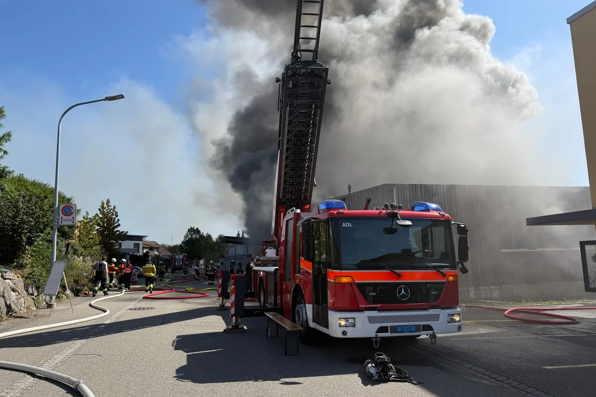 Ein Feuerwehrauto mit ausfahrbarer Leiter steht in einer Straße während dichter Rauch in den Himmel aufsteigt. Feuerwehrleute sind vor Ort.
