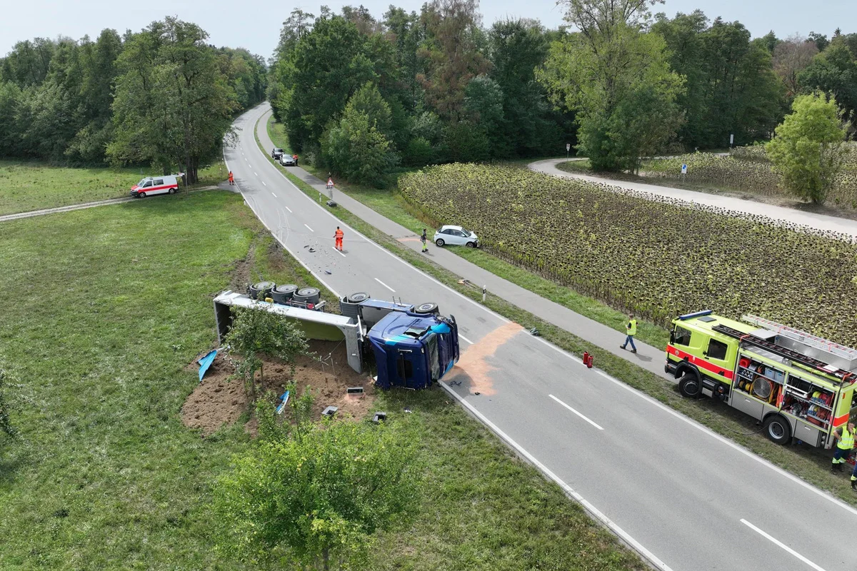 Ein blauer Lkw ist seitlich auf einer Landstraße umgekippt, Erde ist ausgeschüttet. Feuerwehrleute arbeiten vor Ort.