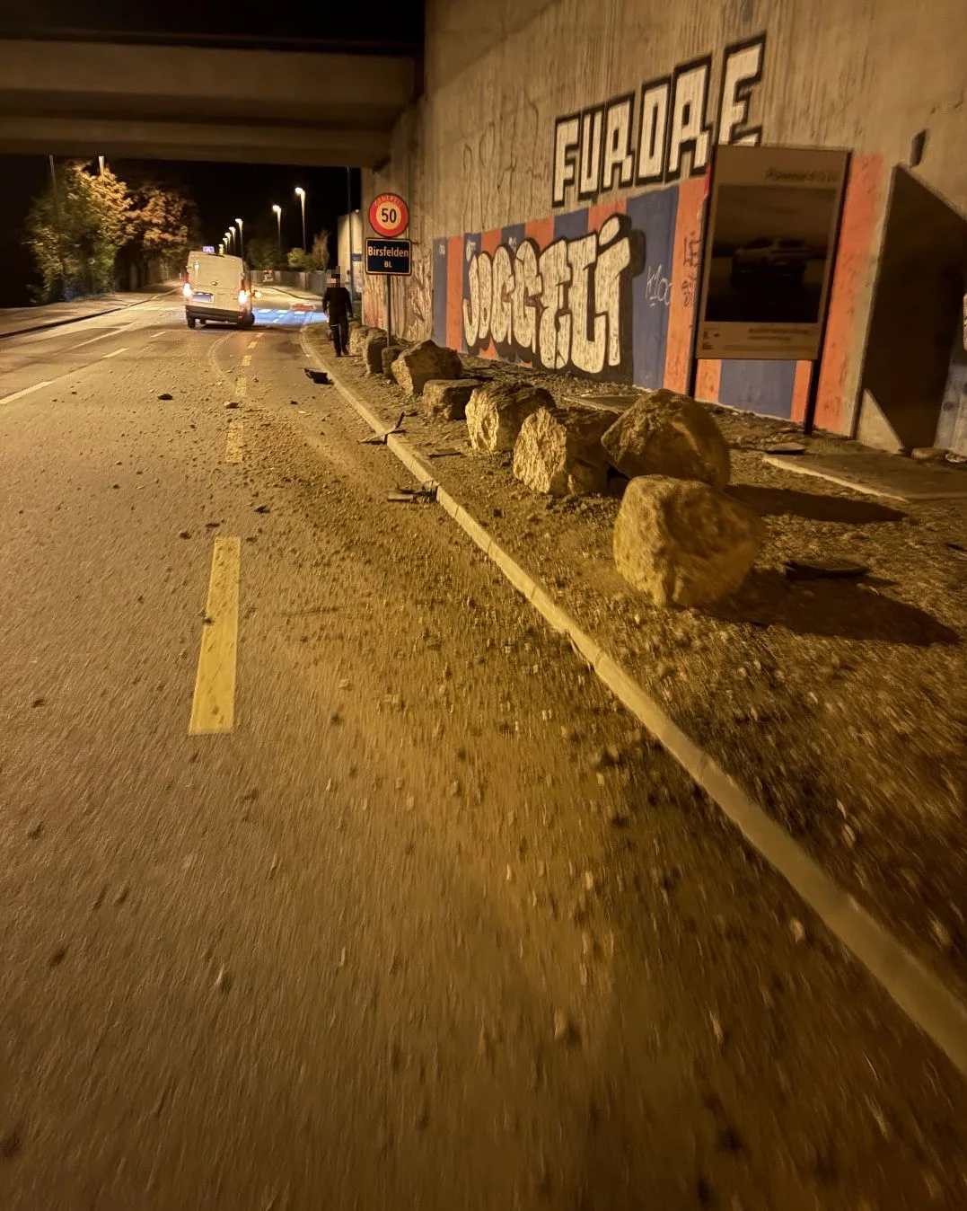 Eine nächtliche Straße unter einer Brücke zeigt verstreute Felsbrocken auf dem Gehweg. Im Hintergrund steht ein Transporter.