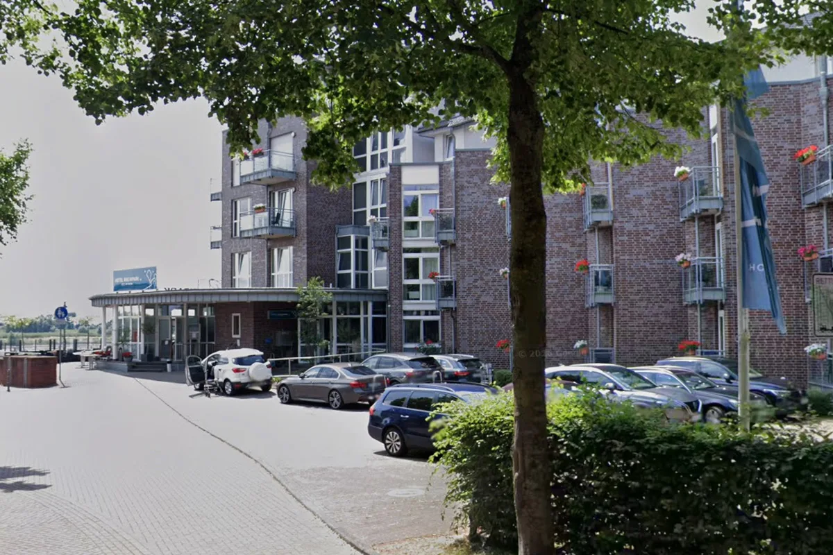 Ein mehrstöckiges Hotel in Ziegelbauweise steht im Hintergrund, davor sind mehrere Autos geparkt. Bäume säumen den Gehweg.