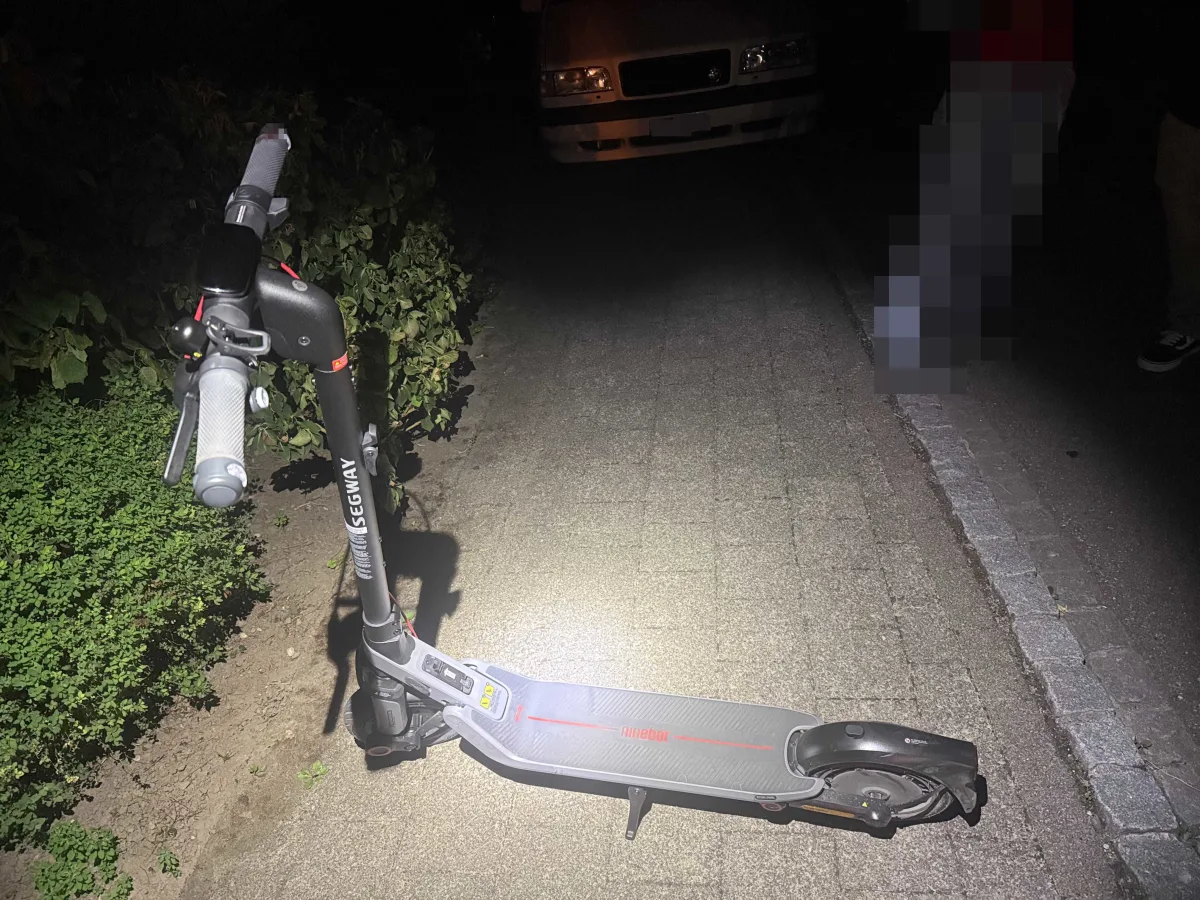 Ein E-Scooter liegt auf einem Gehweg, teilweise von einem Scheinwerfer beleuchtet. Im Hintergrund ist ein geparktes Auto zu sehen.