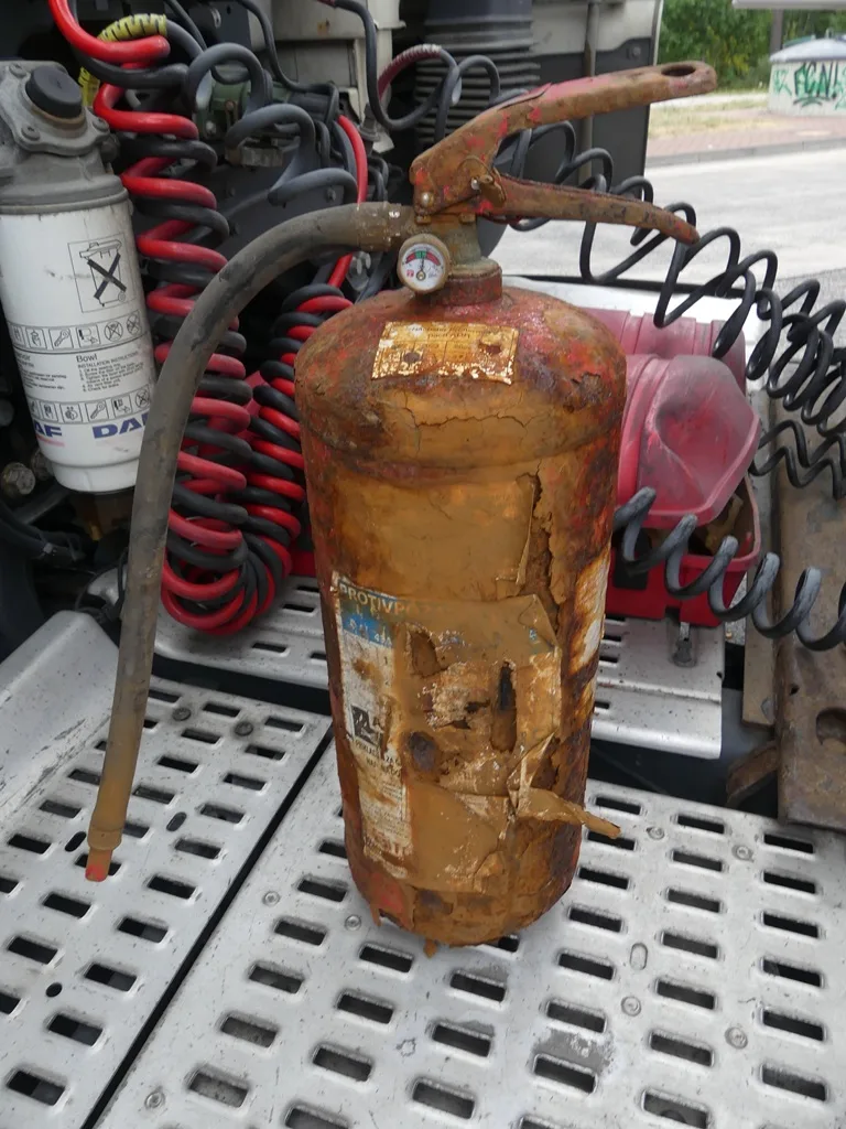 Ein stark rostiger Feuerlöscher steht auf einer Fläche innerhalb eines Fahrzeugs, umgeben von verschiedenen Kabeln und Schläuchen.