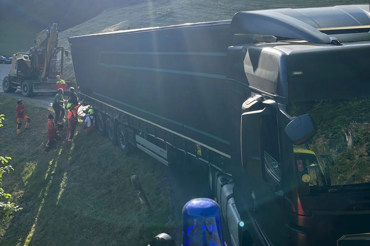 Ein Lastwagen steckt seitlich in einem Graben, während mehrere Personen in Schutzkleidung daran arbeiten. Ein Traktor ist im Hintergrund zu sehen.