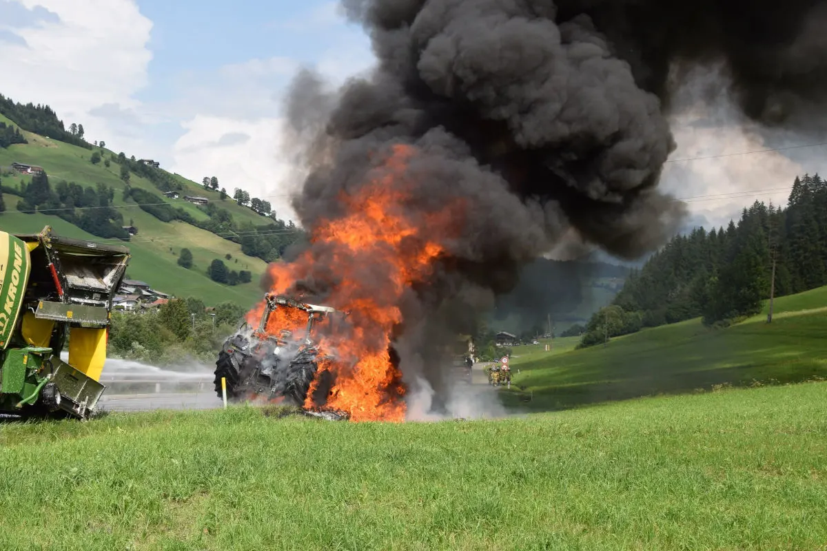 Ein Traktor steht auf einer Wiese in Flammen und erzeugt eine große schwarze Rauchwolke. Im Hintergrund ist eine hügelige Landschaft zu sehen.