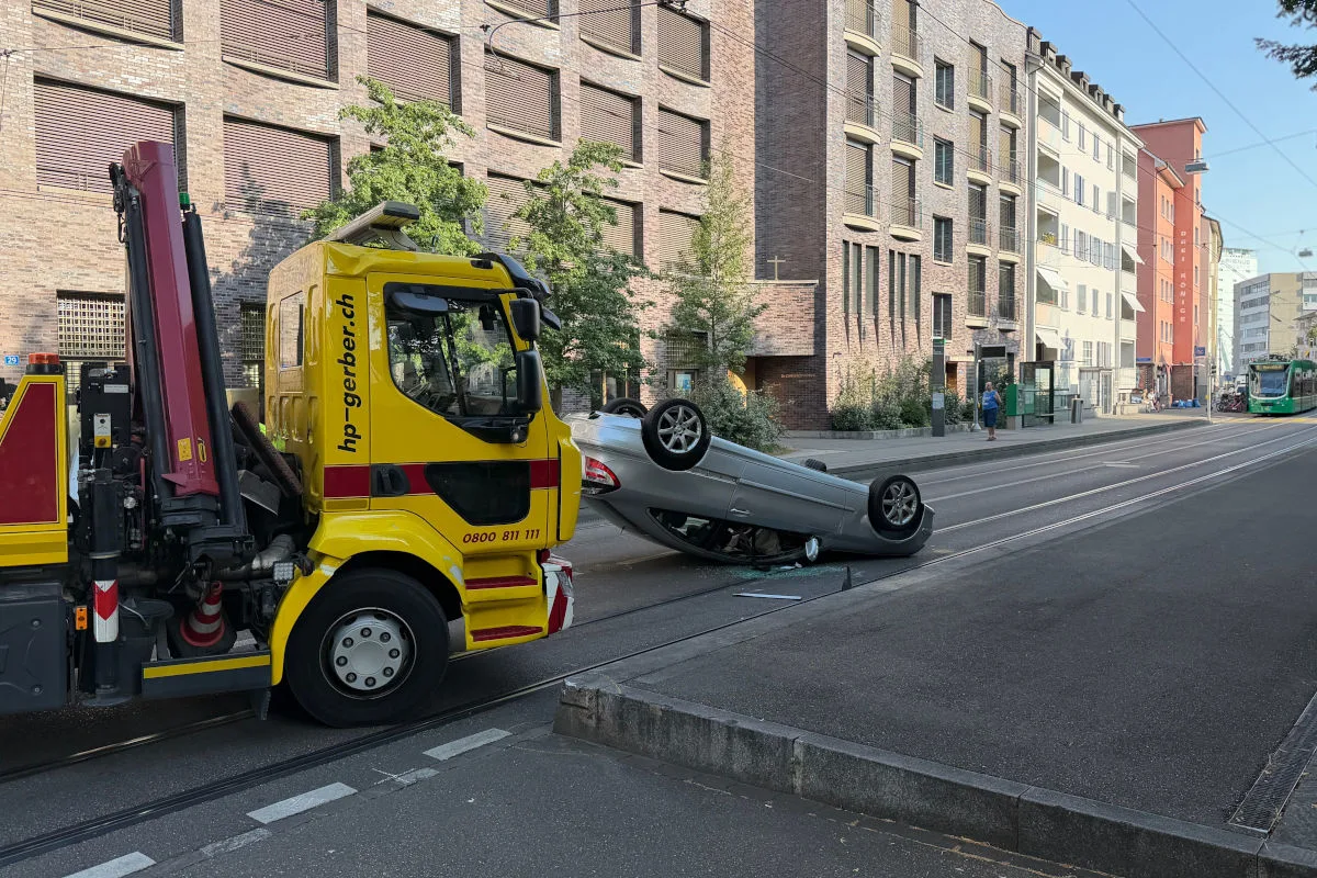 Ein silbernes Auto liegt auf dem Dach auf einer Stadtstraße, neben einem gelben Abschleppfahrzeug. Im Hintergrund sind Wohnhäuser sichtbar.