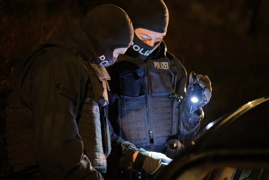 Zwei vermummte Polizisten in taktischer Kleidung inspizieren bei Nacht ein Auto. Sie nutzen Taschenlampen für die Untersuchung.