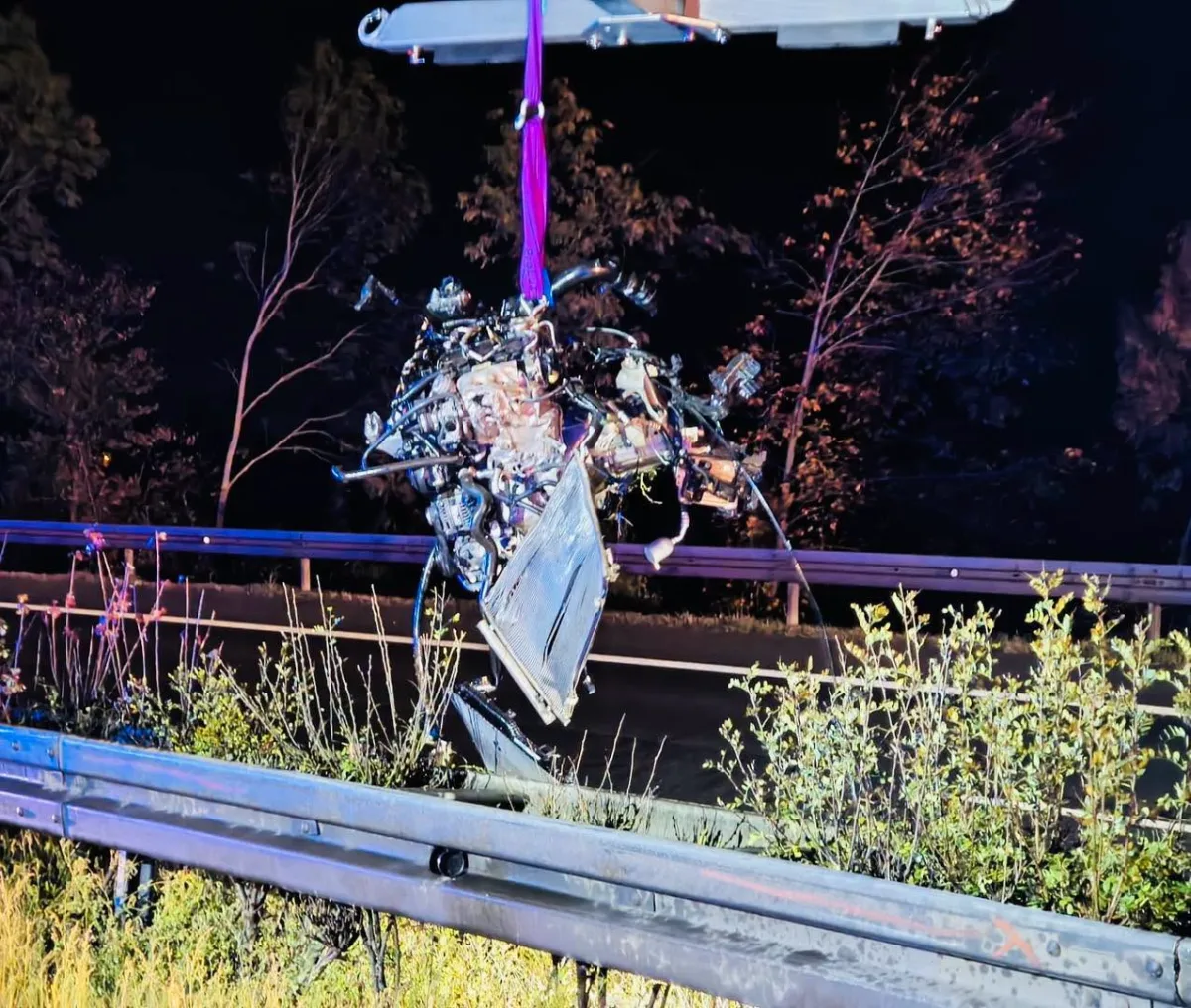 Ein stark beschädigtes Fahrzeugwrack hängt an einem Seil über einer Leitplanke auf einer dunklen Straße. Im Hintergrund sind Bäume sichtbar.