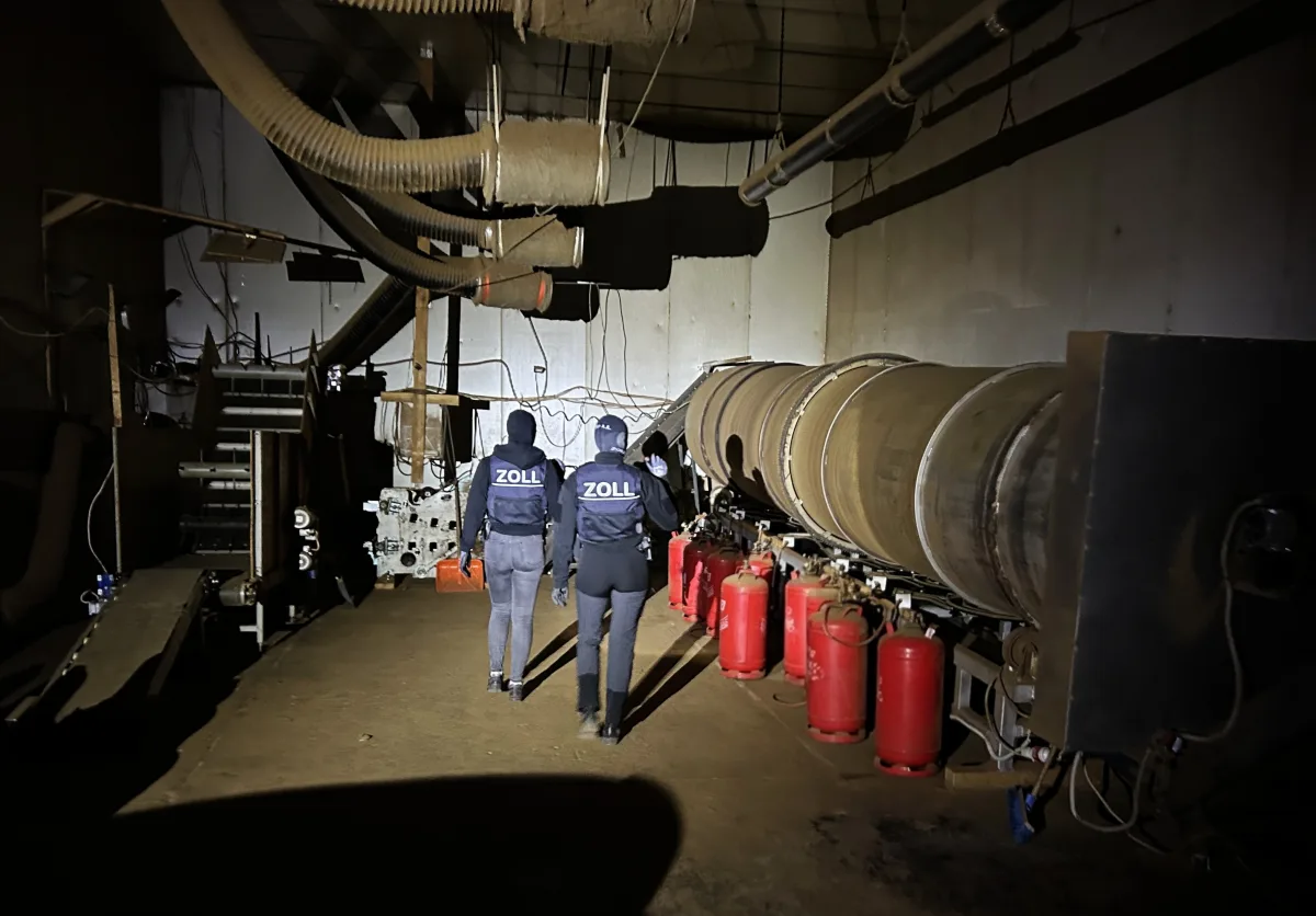 In einer dunkel beleuchteten Halle sind zwei Zollbeamte von hinten zu sehen. Sie tragen Westen mit der Aufschrift 'ZOLL' und inspizieren eine industrielle Anlage mit großen Metallzylindern und roten Gasflaschen.