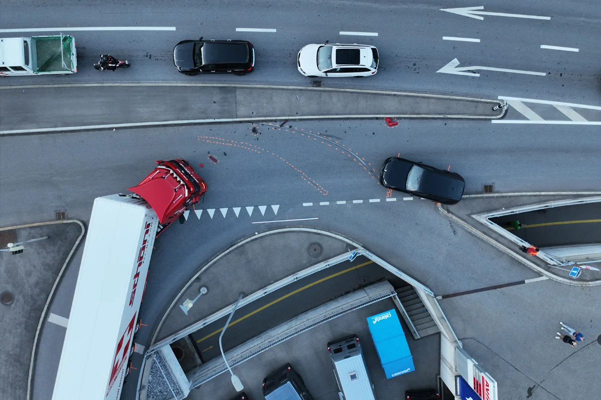 Eine Luftaufnahme zeigt einen Unfall mit einem roten Lkw und mehreren Autos an einer Straße mit Kreuzung, umgeben von urbaner Infrastruktur.