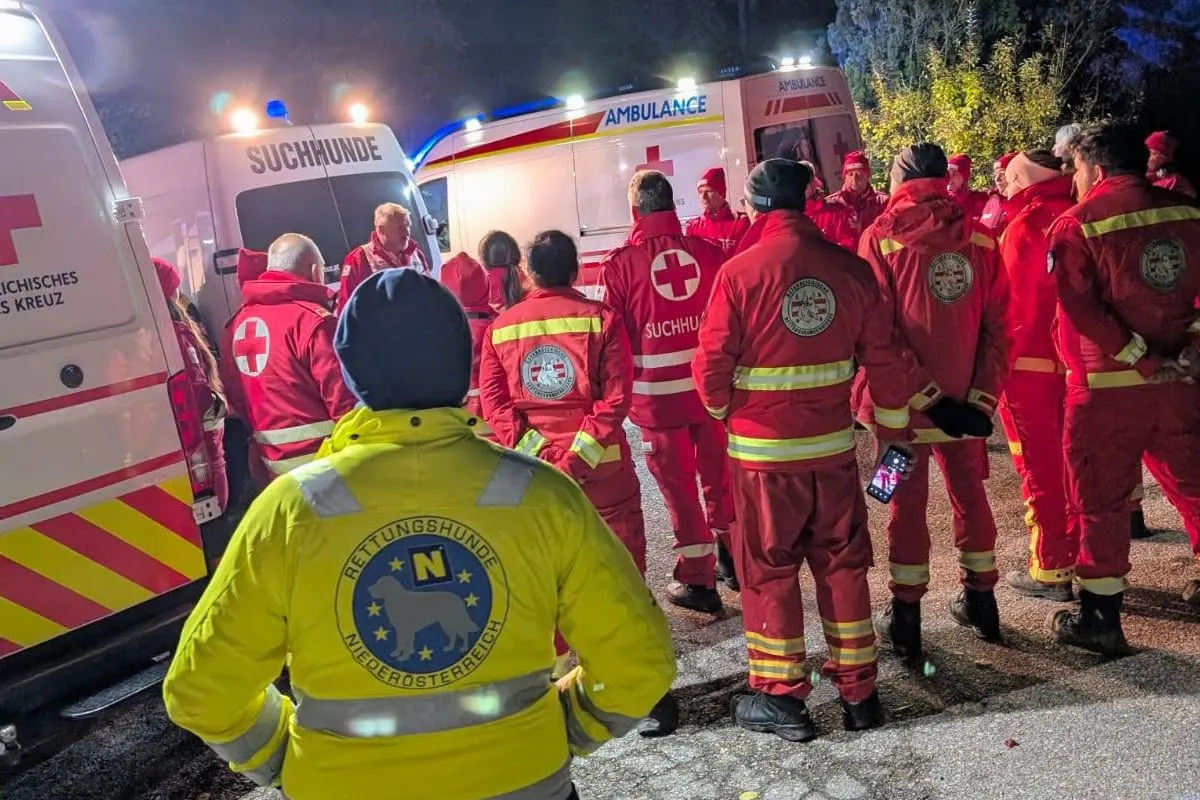 Eine Gruppe von Rettungskräften in roten Uniformen steht vor mehreren Einsatzfahrzeugen. Es ist Nacht, und das Bild wird von Fahrzeuglichtern beleuchtet.