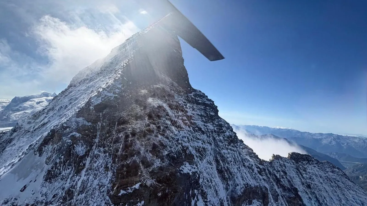 Ein Helikopterrotor ist sichtbar über einem schneebedeckten Berg, umgeben von einer klaren und weiten Berglandschaft. Der Himmel ist blau mit vereinzelten Wolken.