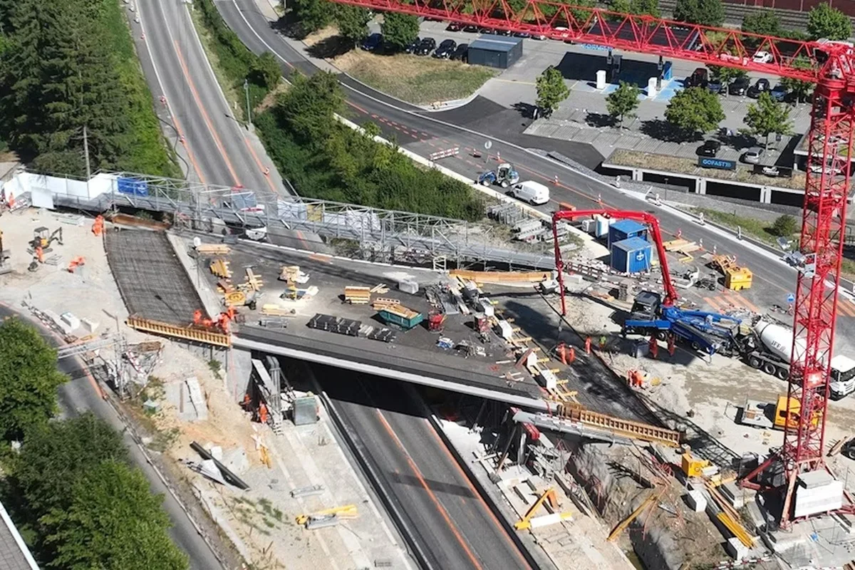 Eine Luftaufnahme zeigt eine Baustelle auf einer Brücke mit Bauarbeitern, einem großen Baukran und verschiedenen Maschinen.
