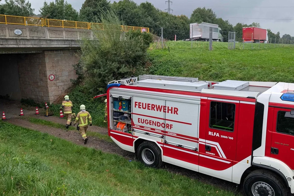 Ein rotes Feuerwehrfahrzeug der Feuerwehr Eugendorf steht nahe einem Tunnel. Zwei Feuerwehrleute in gelben Anzügen gehen darauf zu.