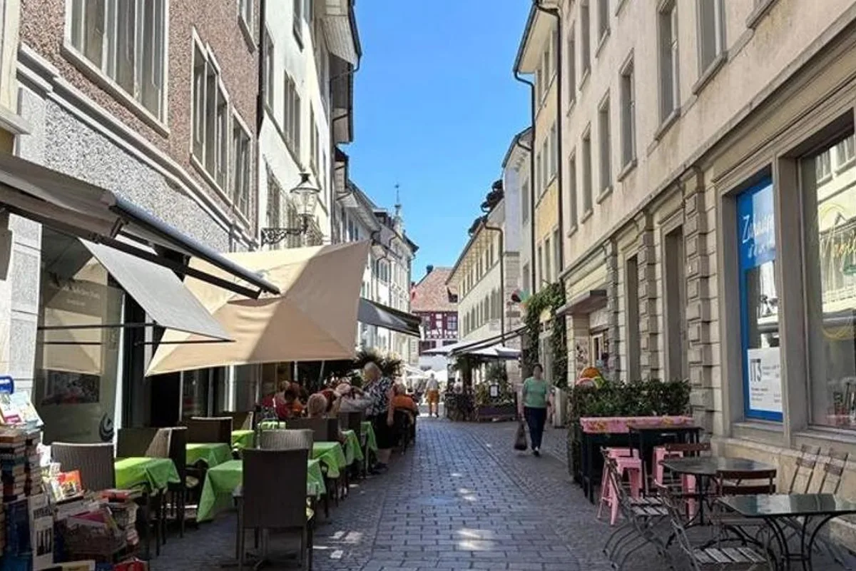 In einer engen, gepflasterten Gasse einer Altstadt sitzen Menschen an Tischen eines Straßencafés. Die Szene ist hell beleuchtet durch sonniges Wetter.