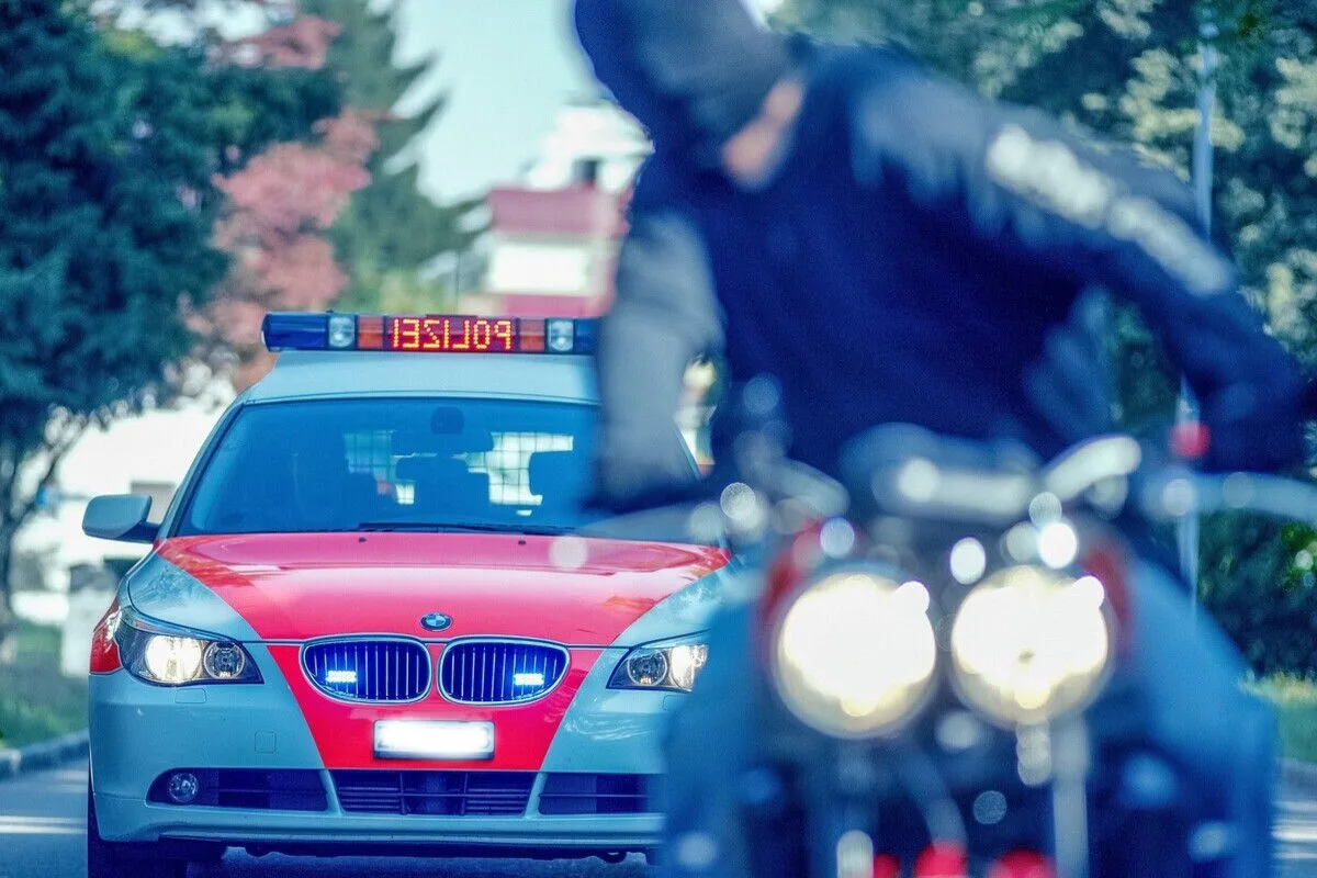 Ein Polizeiwagen mit eingeschaltetem Blaulicht fährt hinter einem Motorradfahrer her. Der Polizeiwagen hat eine rote und blaue Lackierung.