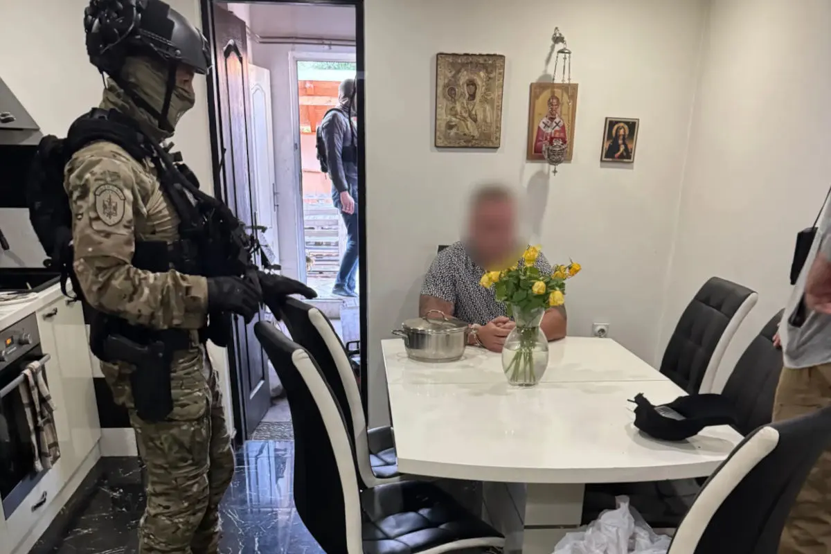 Ein Polizist in taktischer Ausrüstung steht in einer Küche, während eine Person am Tisch sitzt. Ein weiterer Polizist steht im Türbereich.