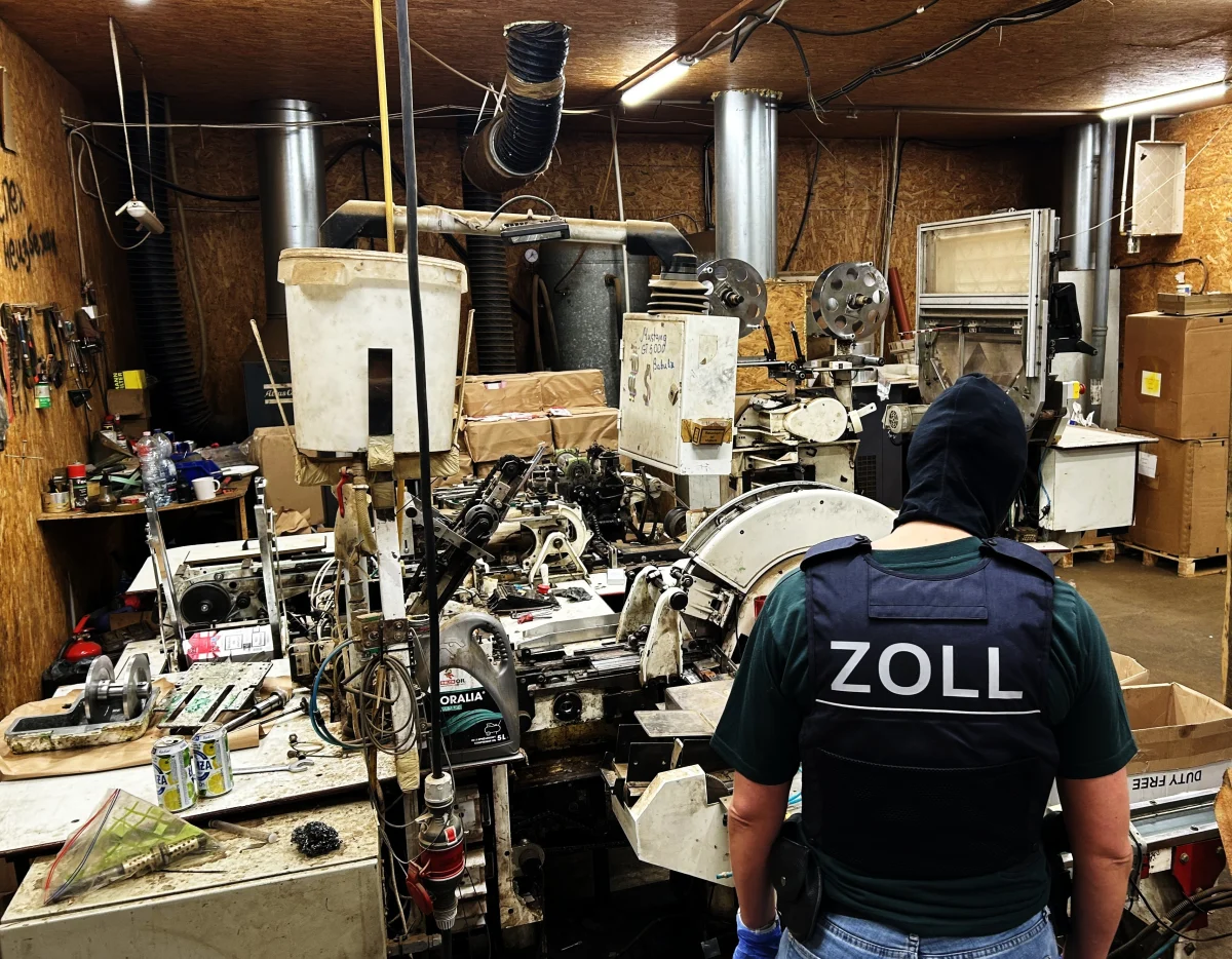 Ein Zollbeamter in Uniform inspiziert eine Werkstatt mit komplexen Maschinen und vielen Kartons. Die Szene deutet auf eine illegale Produktionsstätte hin.