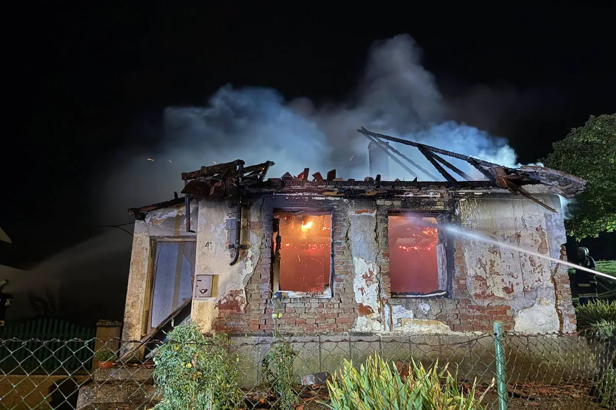 Das Bild zeigt ein stark beschädigtes Haus bei Nacht mit Flammen im Inneren und Rauchentwicklung. Feuerwehreinsatzkräfte löschen den Brand.