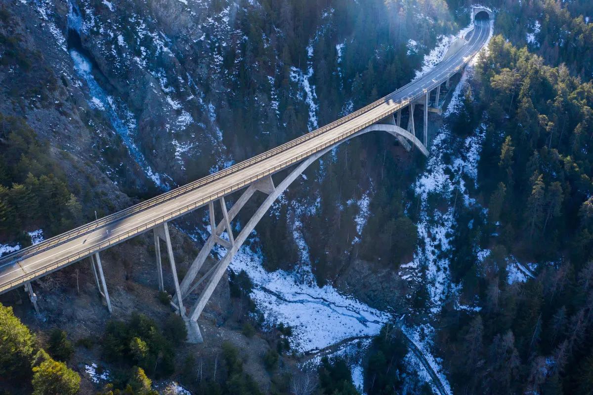 Die Viamala-Schluchtbrücke erstreckt sich über eine tiefe, schneebedeckte Schlucht mit bewaldeten Hängen. Am Ende der Brücke führt ein Tunnel in den Berg.
