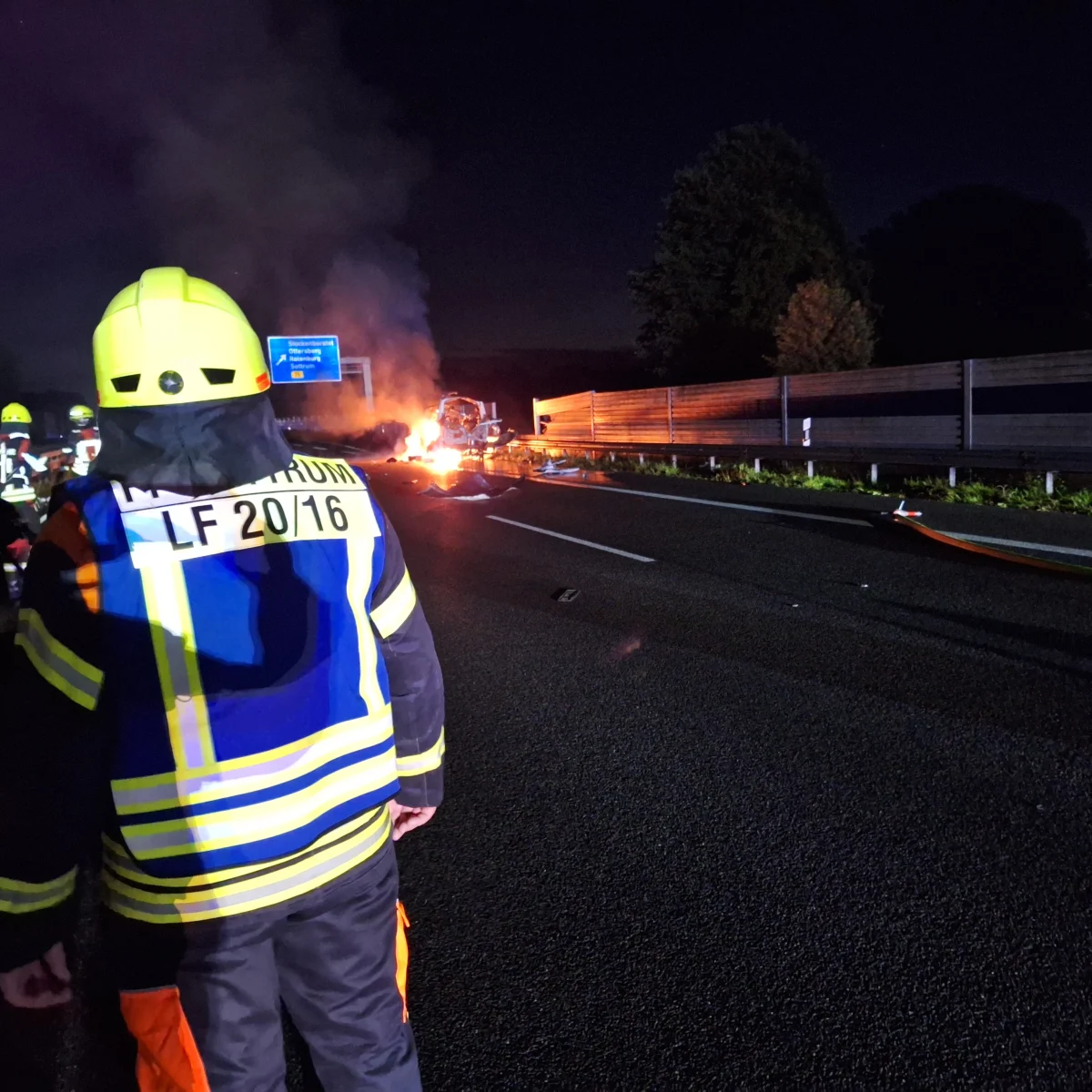 Ein Feuerwehrmann im Vordergrund schaut auf ein brennendes Fahrzeug auf einer nächtlichen Autobahn. Rauch steigt aus dem Fahrzeug auf.