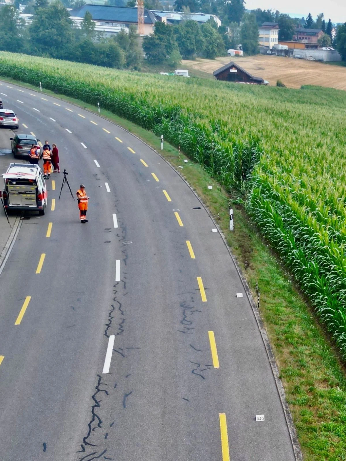 Ein Einsatzfahrzeug parkt auf einer Landstraße, während mehrere Personen in orangefarbener Kleidung Spuren sichern. Ein Maisfeld ist daneben sichtbar.