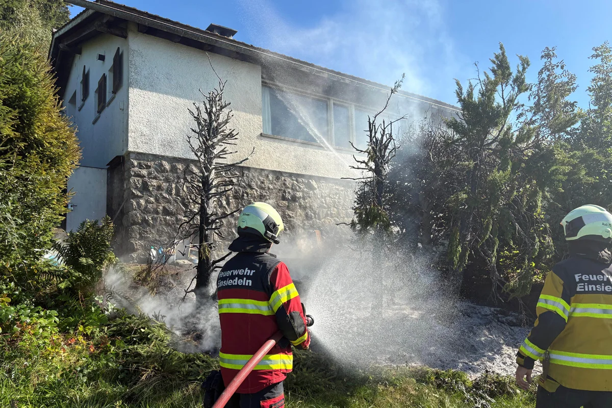 Zwei Feuerwehrleute der Feuerwehr Einsiedeln löschen mit einem Schlauch ein Feuer neben einem Gebäude; verkohlte Bäume sind im Vordergrund sichtbar.