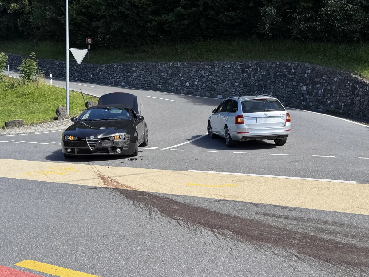 An einer Straßenkurve stehen zwei Autos, ein schwarzer Alfa Romeo mit geöffneter Motorhaube und Frontschaden und ein weißer Kombi.