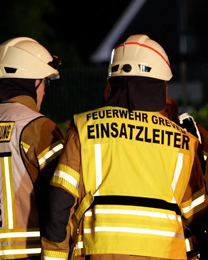 Zwei Feuerwehrleute in gelben Westen und Helmen stehen nebeneinander. 'Einsatzleiter' ist auf der Weste sichtbar.