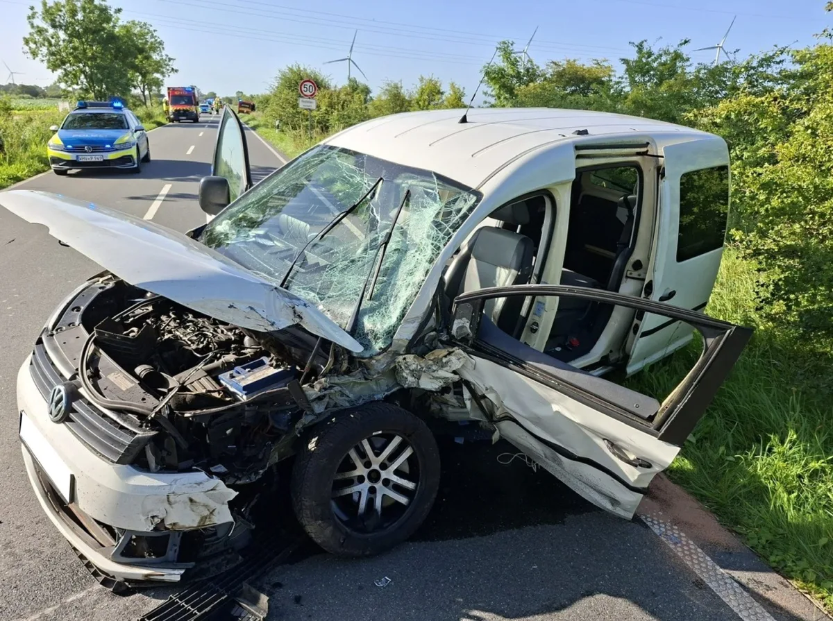 Ein stark beschädigter weißer VW mit offenem Motorraum steht auf einer Landstraße. Polizei- und Feuerwehrfahrzeuge sind im Hintergrund sichtbar.