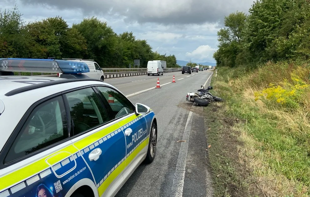 Ein Polizei-Einsatzfahrzeug steht auf dem Seitenstreifen einer Autobahn, neben einem umgestürzten Motorrad auf der Grasfläche. Der Verkehr fließt weiter auf der Fahrbahn.
