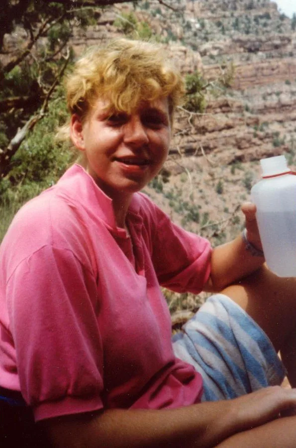 Eine lächelnde Person in einem pinken T-Shirt sitzt im Freien und hält eine Plastikflasche. Im Hintergrund ist eine felsige Landschaft zu sehen.