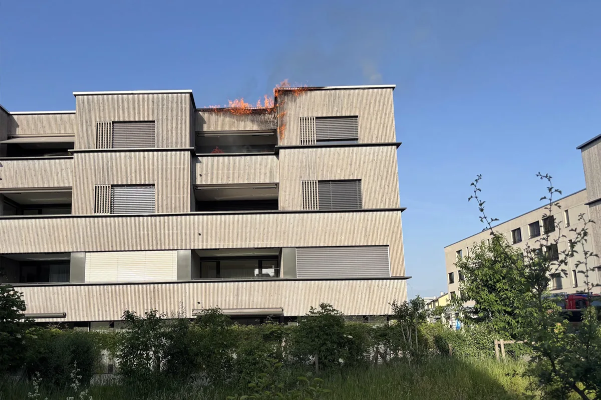 Ein modernes Mehrfamilienhaus mit Flammen am Dachrand; Rauch steigt auf.