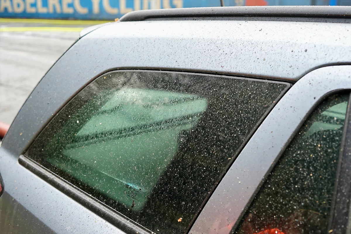 Ein geparktes Auto im Freien mit schmutzigen Fensterscheiben voller Sand- und Ascherückstände. Im Hintergrund ist eine Absperrung und ein Schild sichtbar.
