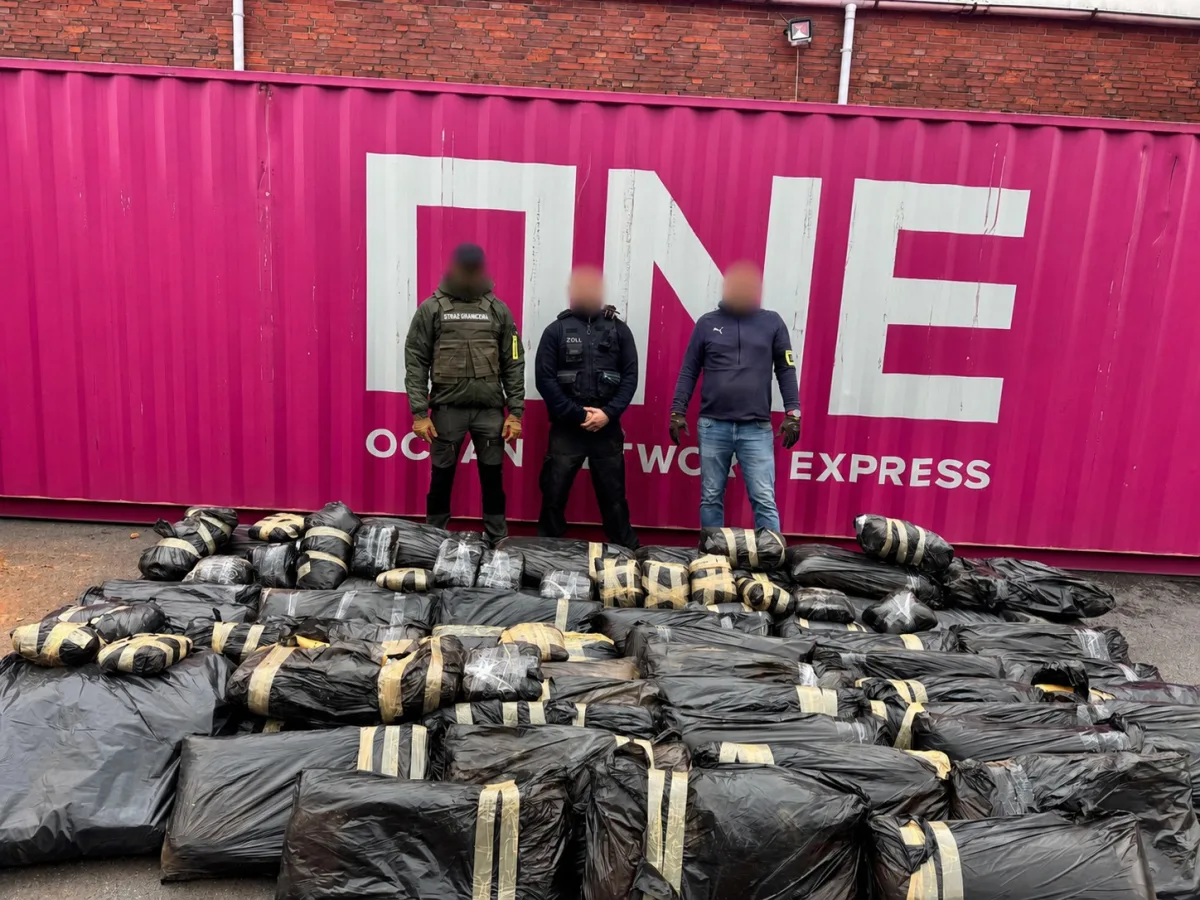 Drei Personen stehen vor einem rosa Container, auf dem Boden liegen zahlreiche in Plastik verpackte Pakete gestapelt.