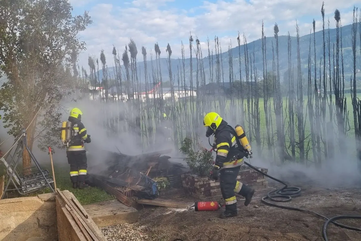 Zwei Feuerwehrleute in gelben Helmen und Atemschutzgeräten löschen ein Feuer auf einer Wiese mit Schläuchen und Löschwasser.