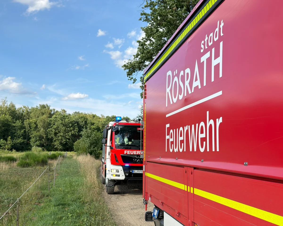 Zwei Feuerwehrfahrzeuge der Stadt Rösrath stehen auf einem Feldweg neben einem bewaldeten Gebiet. Der Himmel ist klar mit wenigen Wolken.