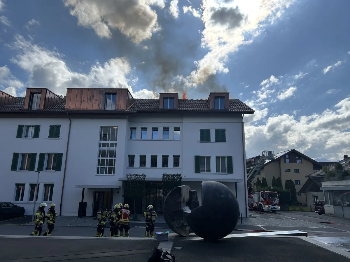 Ein mehrstöckiges Wohngebäude zeigt Rauch und Flammen, die aus dem Dach kommen. Mehrere Feuerwehrleute stehen vor dem Gebäude, ein Feuerwehrfahrzeug ist am rechten Bildrand zu sehen.