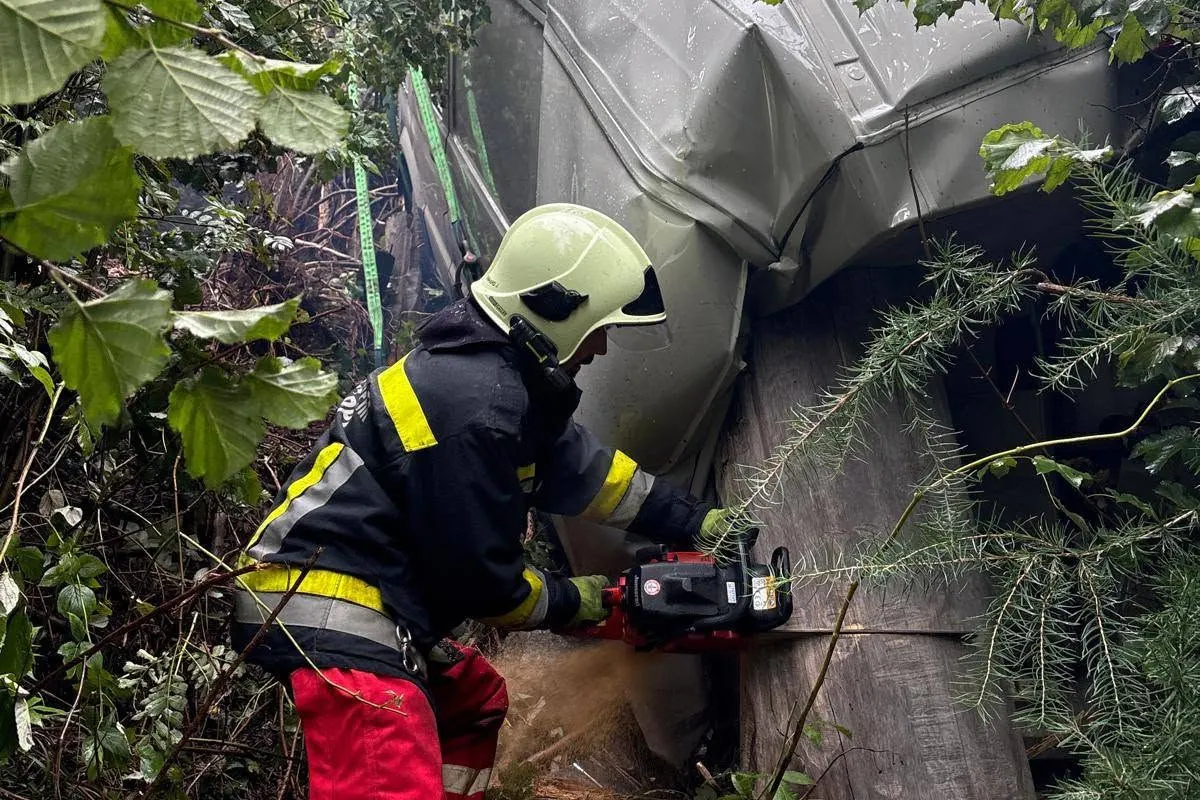 Ein Feuerwehrmann trägt Schutzkleidung und nutzt eine Kettensäge, um Äste von einem verunfallten Fahrzeug zu entfernen. Der Unfall ereignete sich in einer bewaldeten Umgebung.
