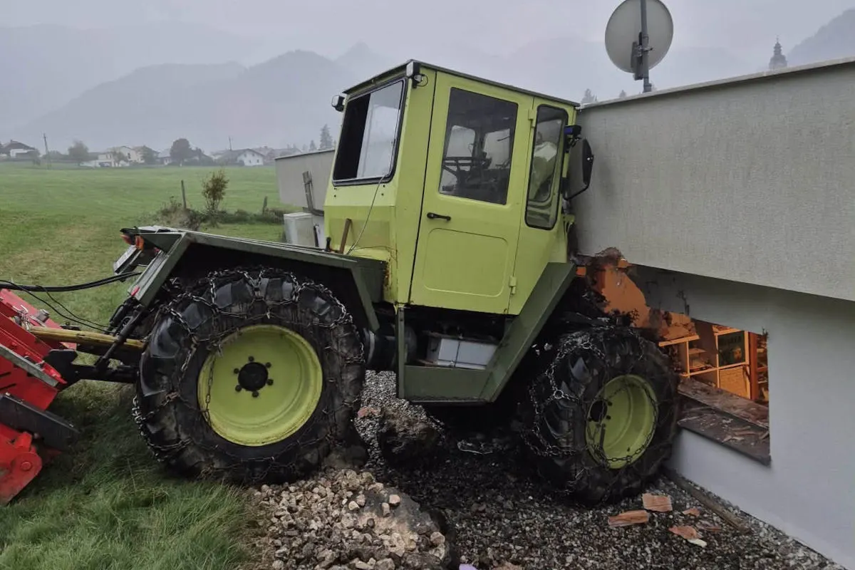 Ein grüner Traktor ist frontal mit einer Hauswand kollidiert, wodurch ein Loch in der Fassade entstanden ist. Es ist bewölkt und die Umgebung ist ländlich mit Wiesen und wenigen Gebäuden.
