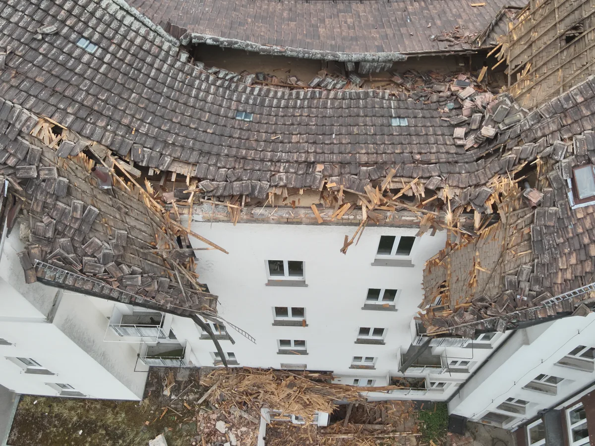 Das Bild zeigt ein mehrstöckiges Gebäude mit einem stark beschädigten Dach, das teilweise eingestürzt ist. Holzteile und Ziegel sind lose und zersplittert, einige Teile liegen am Boden.