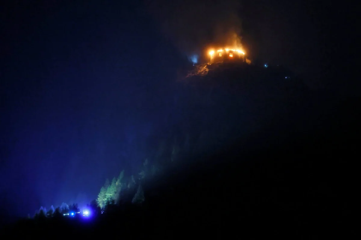 In der Nacht brennt ein Gebäude auf einem Hügel, Flammen sind deutlich sichtbar. Unten leuchten Lichter der Einsatzkräfte.