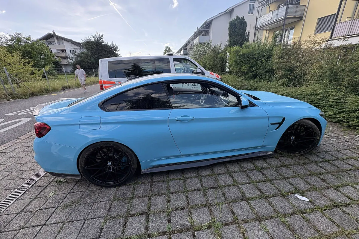 Ein blaues Auto ist auf einem gepflasterten Weg in einer Wohngegend geparkt. Im Hintergrund steht ein Polizeifahrzeug, ein Mann geht auf dem Gehweg.