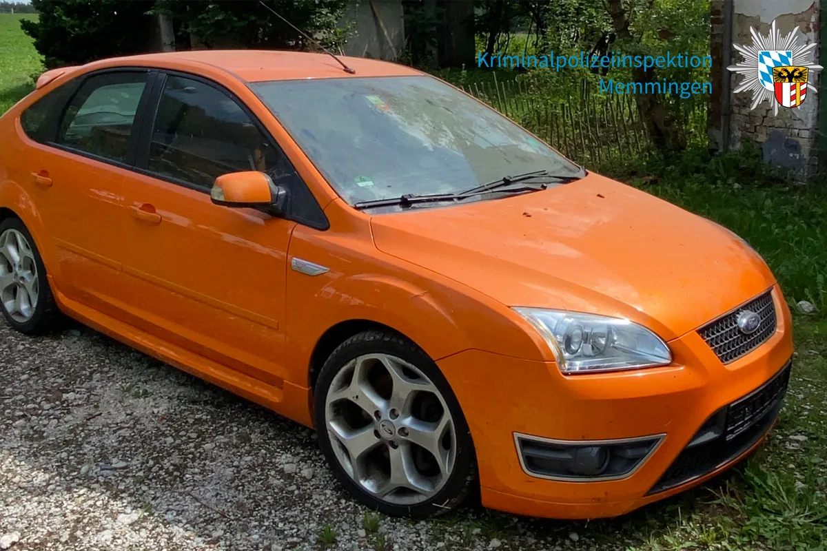 Ein orangefarbener Ford steht auf einem Schotterweg. Im Hintergrund ist ein älteres Gebäude und Vegetation erkennbar.