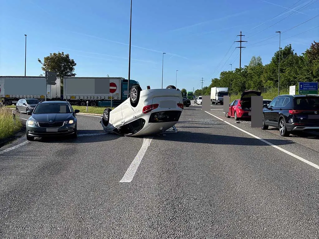 Ein umgekippter, weißer PKW liegt auf dem Dach auf einer Fahrbahn, umgeben von weiteren Autos bei Tageslicht. LKWs und Autos sind in der Nähe.