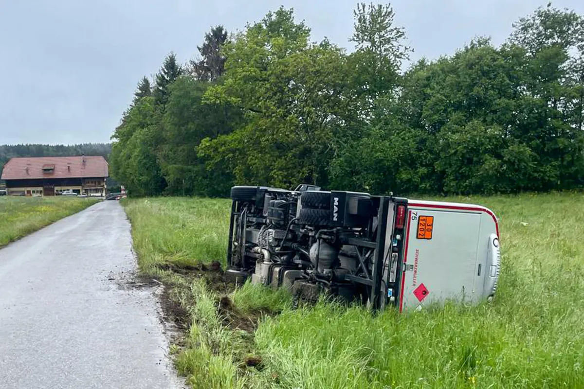 Ein Lastwagen liegt auf der Seite in einer Wiese neben einer schmalen, ländlichen Straße. Im Hintergrund sind Bäume und ein Hof zu sehen.