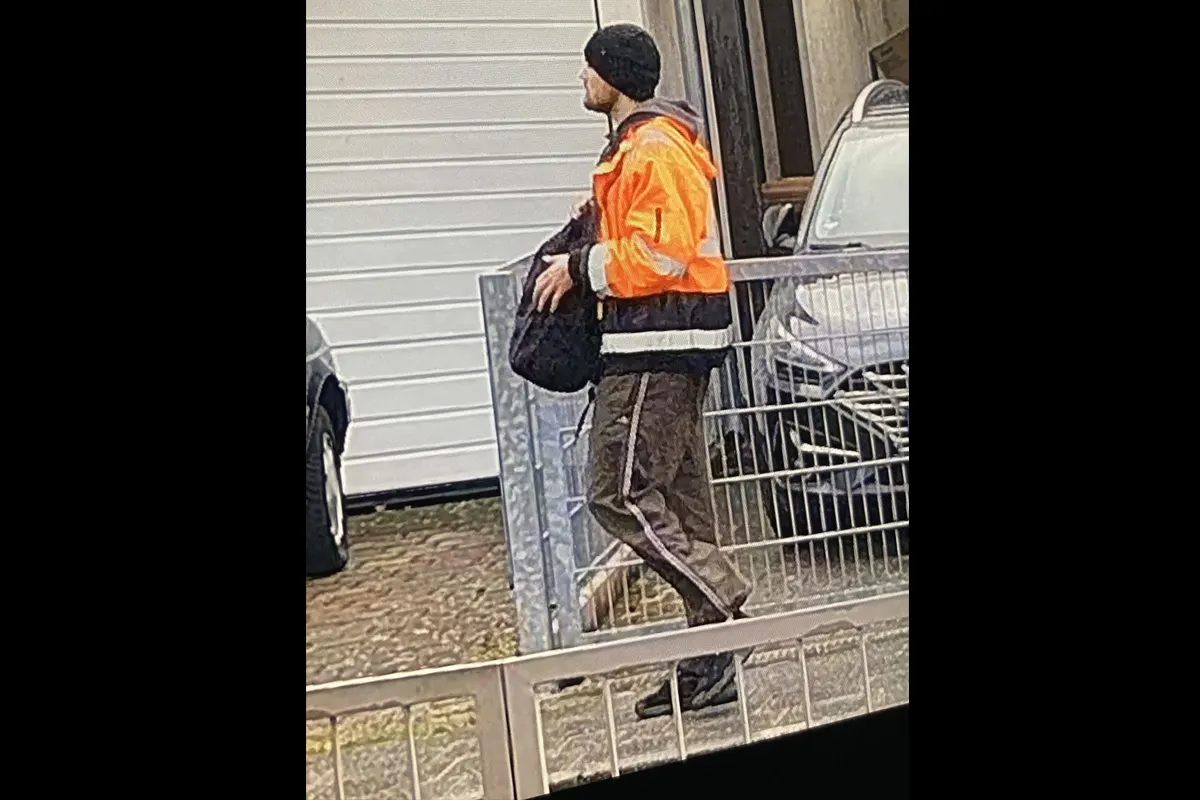 Ein Mann in einer leuchtend orangefarbenen Jacke und einem schwarzen Rucksack steht neben einem geparkten Auto hinter einem Metallzaun. Die Szene scheint in einem Wohn- oder Garagenbereich aufgenommen worden zu sein.