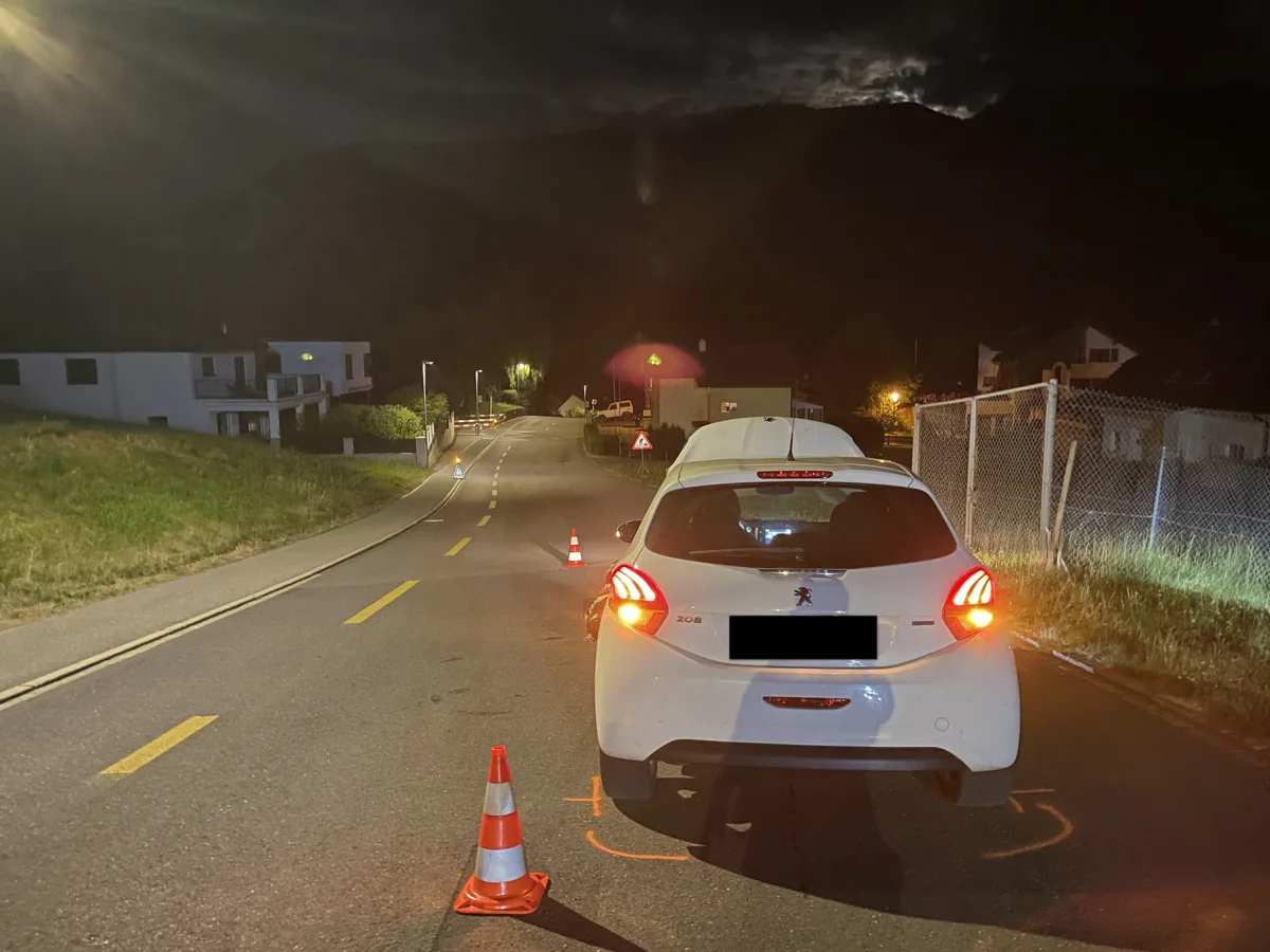 Ein weißes Auto ist auf einer Landstraße bei Nacht, umgeben von mehreren orangefarbenen Verkehrskegeln zu sehen. Häuser und Straßenbeleuchtung sind im Hintergrund sichtbar.