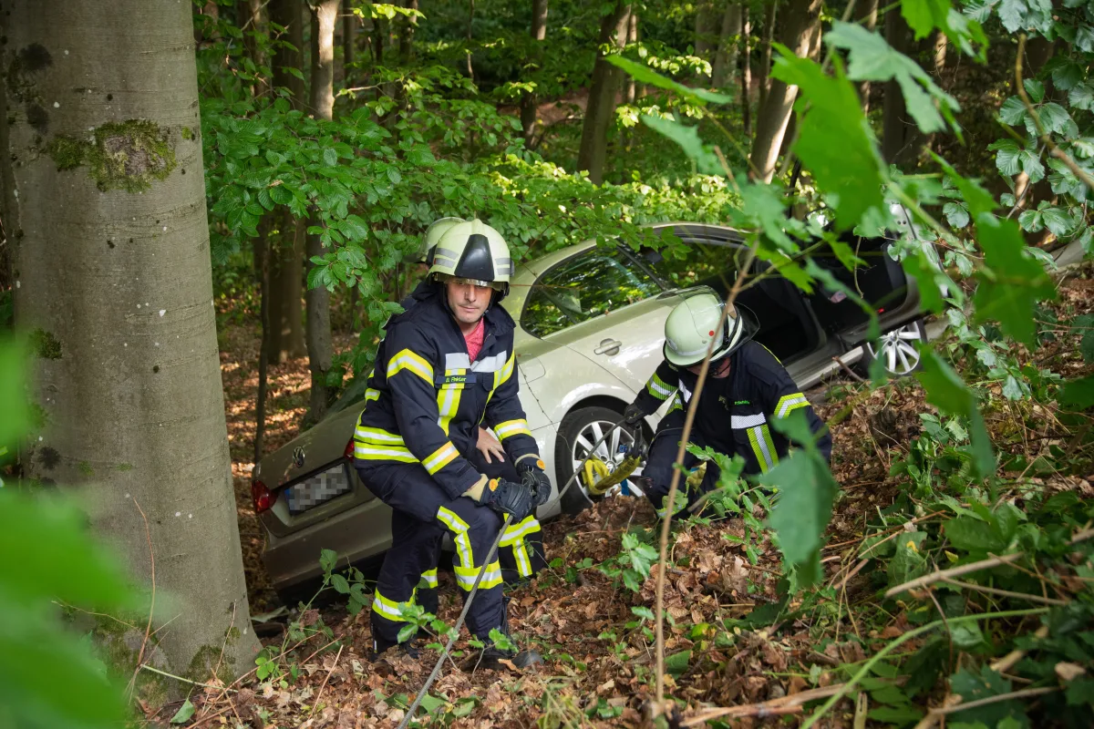 Zwei Feuerwehrleute arbeiten an einem Auto, das in einem Waldstück von der Straße abgekommen ist. Das Auto steht teilweise verdeckt durch Bäume und Laub, während die Feuerwehrleute es sichern.