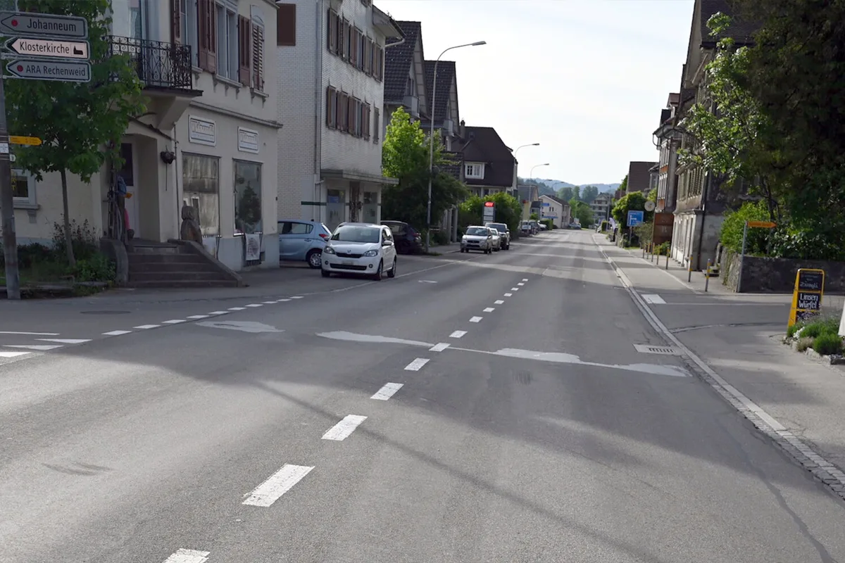 Eine leere Straße in einem Dorf, flankiert von Gebäuden und geparkten Autos. Einige Wegweiser sind sichtbar.