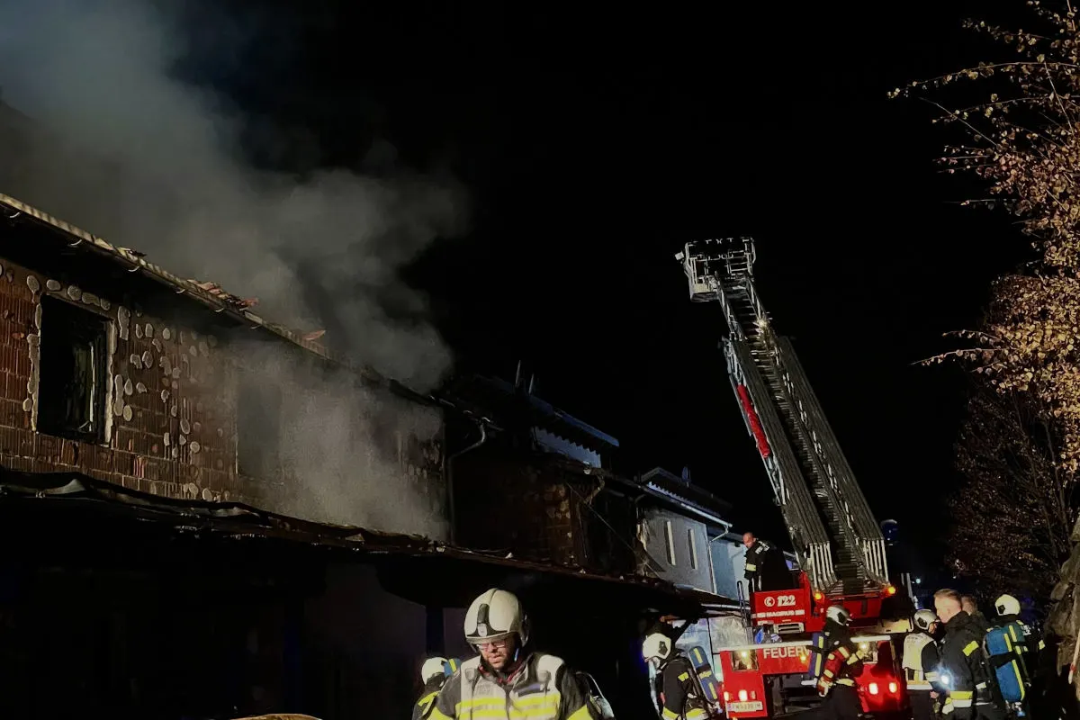 Mehrere Feuerwehrleute sind im Einsatz, um ein brennendes Haus in der Nacht zu löschen. Eine Drehleiter ist in Aktion, während Rauch aus dem Gebäude aufsteigt.